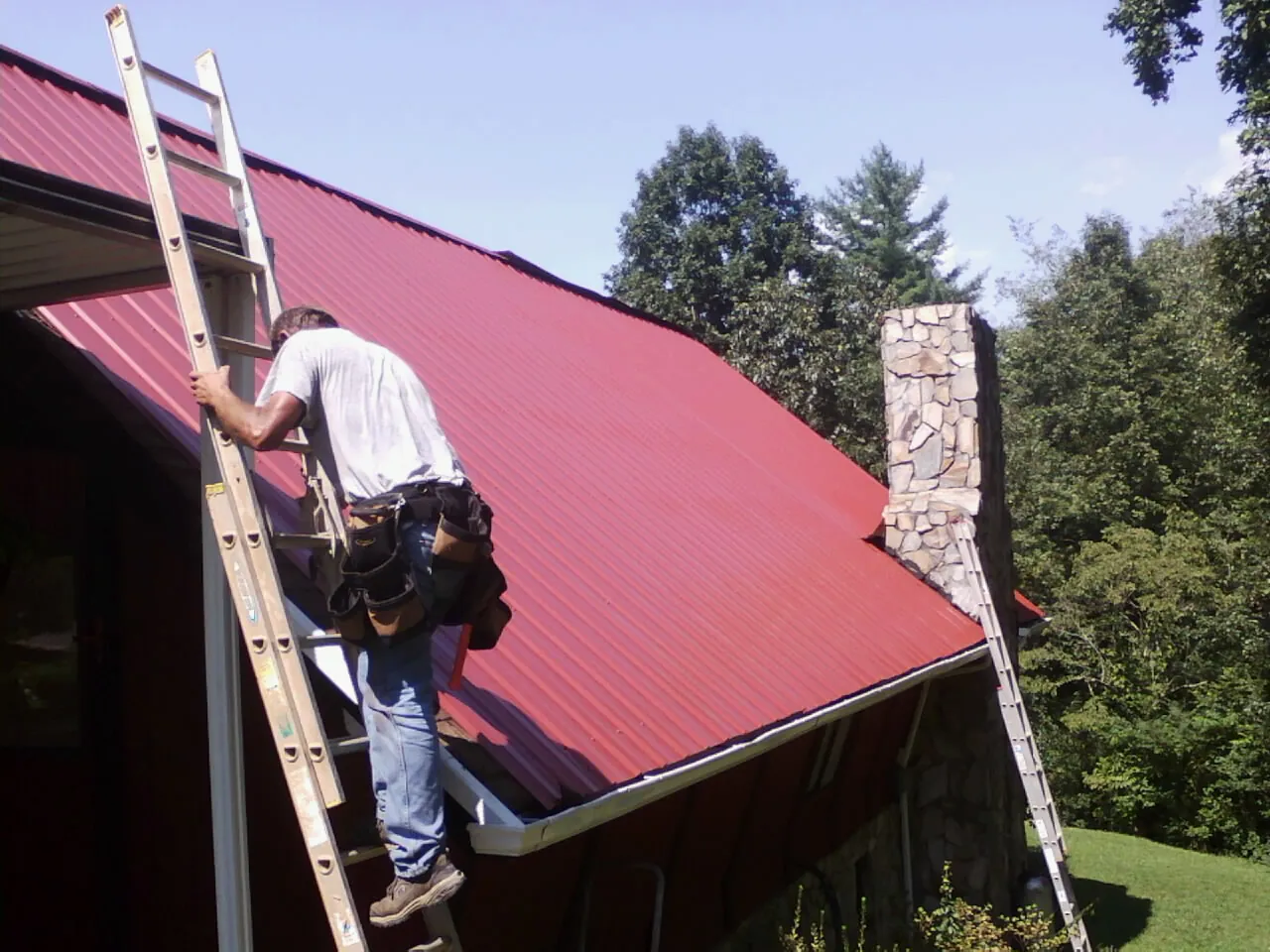 Roofer on a ladder beside a red metal roof and stone chimney — Soffit & Fascia Repair by Roofing Brothers in Palacios